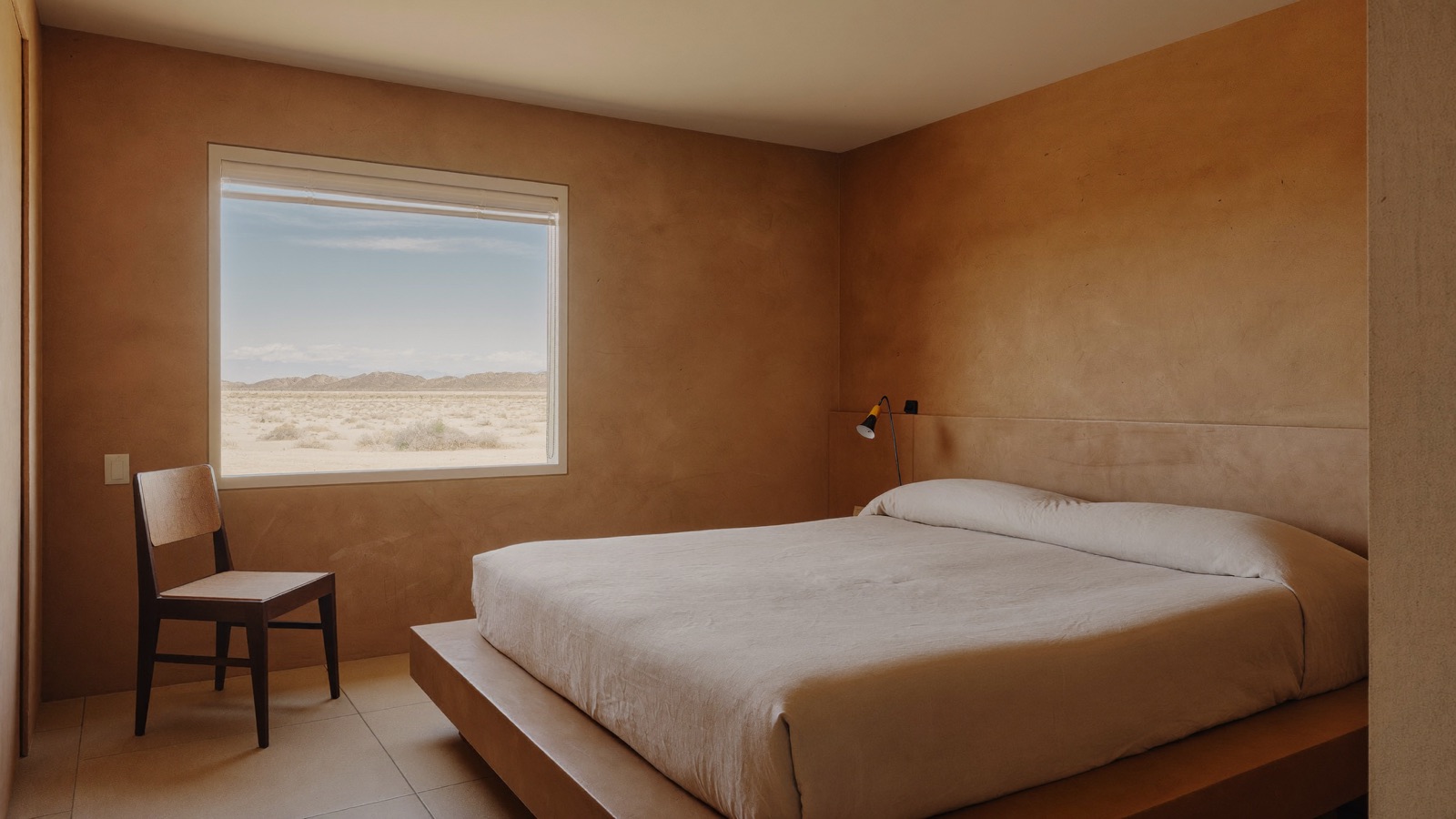 A room at Sonora House at dawn: a minimalist adobe interior with a linen-dressed bed and a window framing the desert mesas in soft morning light